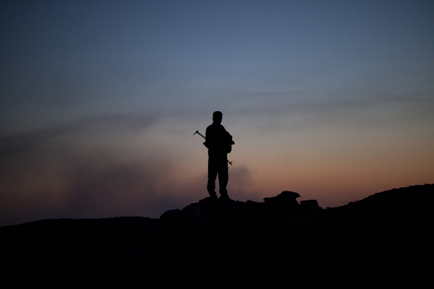 Un Peshmerga controla un posible ataque del Estado Islámico en uno de los puntos estratégicos de la línea de Tuz Khurmatu. Al fondo, a la luz del crepúsculo, el humo refleja la actividad de los miembros del Estado Islámico. Tuz Khurmatu; Kurdistán, Irak. 19 de junio de 2017