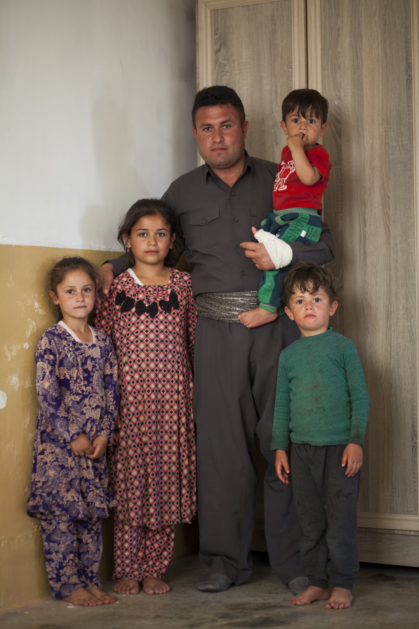 Abdulla (34), former Peshmerga, poses before the camera with his family. A bullet went through his neck in 2016 while fighting the IS in Kirkuk. After being resucitated he remained unconscious for five days in hospital, and spent months unable to talk, see or eat. The bullet caused paralysis. Currently (2017), he has been in bed for 7 months requiring constant care. He did not fight for any political party, but to see his children grow up. Ranya, Kurdistan, Iraq. May 14, 2017